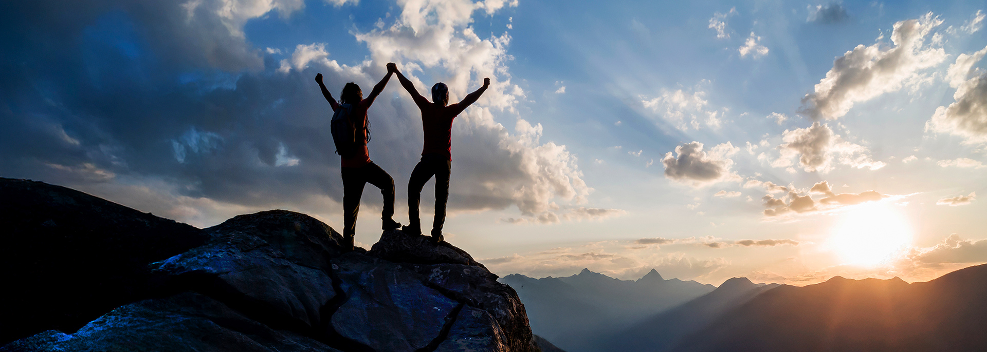Zwei Personen mit Rucksäcken stehen auf einem Felsen und halten sich an den Händen vor einem Bergpanorama bei Sonnenuntergang