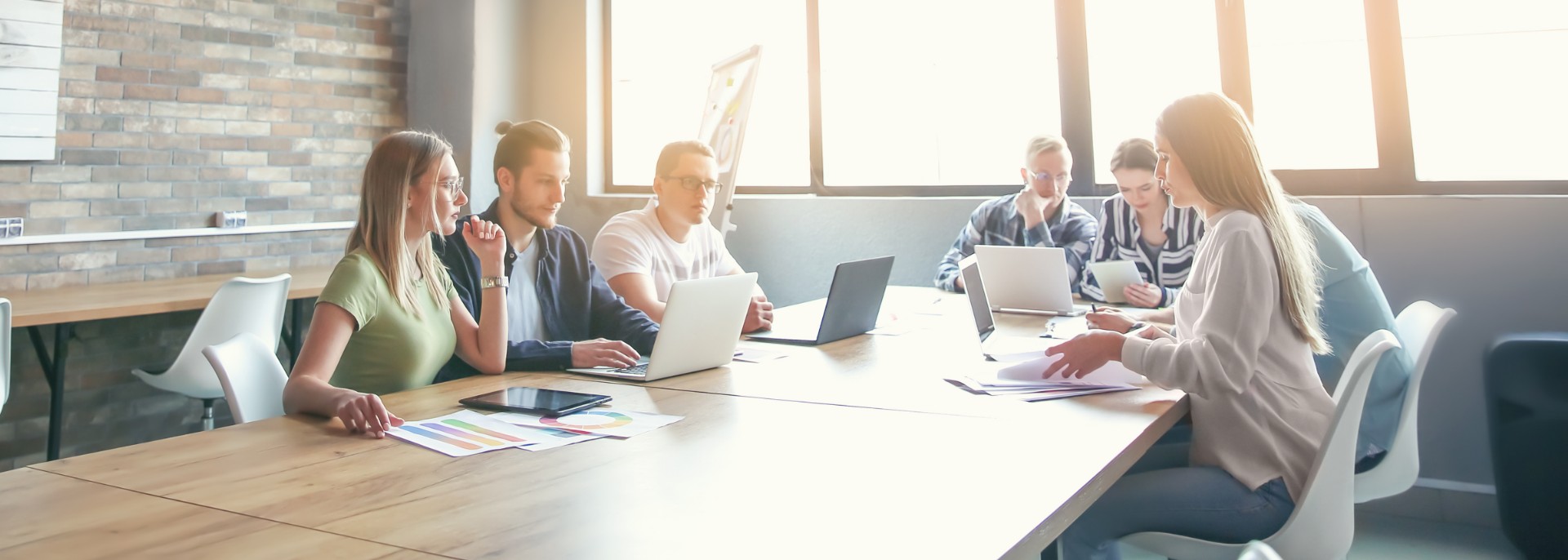Gruppe von sechs Personen sitzt an einem langen Holztisch in einem modernen, hellen Büro mit großen Fenstern und Ziegelwand und arbeitet gemeinsam an Laptops und Dokumenten